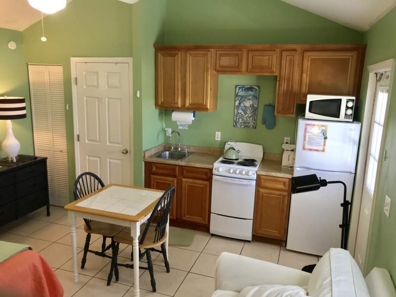 Light green kitchenette with oak cabinets, small stove, microwave, refrigerator, two person table, partial image of couch and bed
